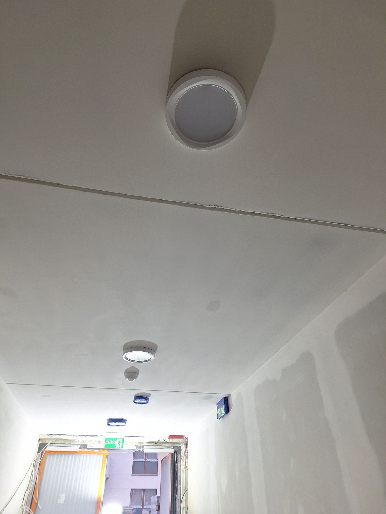 Biały sufit korytarza z zamontowanymi okrągłymi lampami sufitowymi LED i oznaczeniami ewakuacyjnymi. Widoczne ślady po szpachlowaniu.