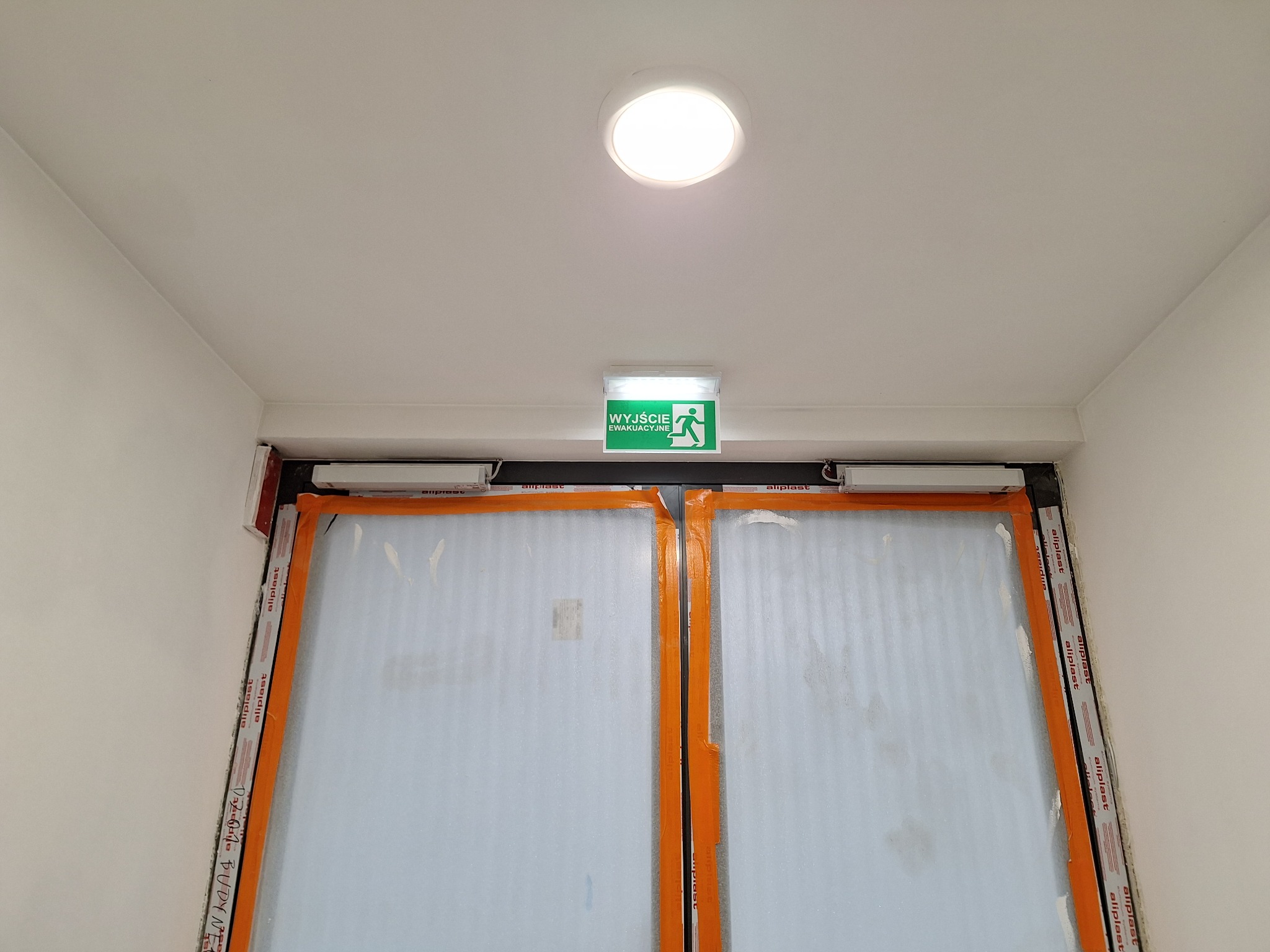 Białe drzwi ewakuacyjne z pomarańczowym zabezpieczeniem, oświetlone okrągłą lampą sufitową i podświetlonym znakiem 'Wyjście Ewakuacyjne' na białym suficie.