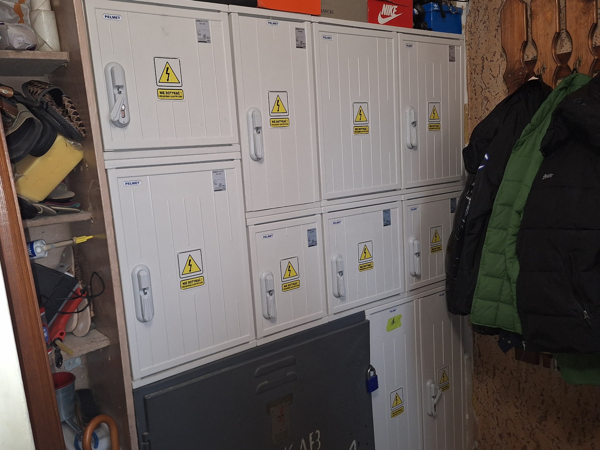 Zestaw białych szafek elektrycznych PELMET z ostrzeżeniami 'Nie dotykać' w pomieszczeniu gospodarczym, obok regału z przedmiotami i wiszącymi kurtkami.