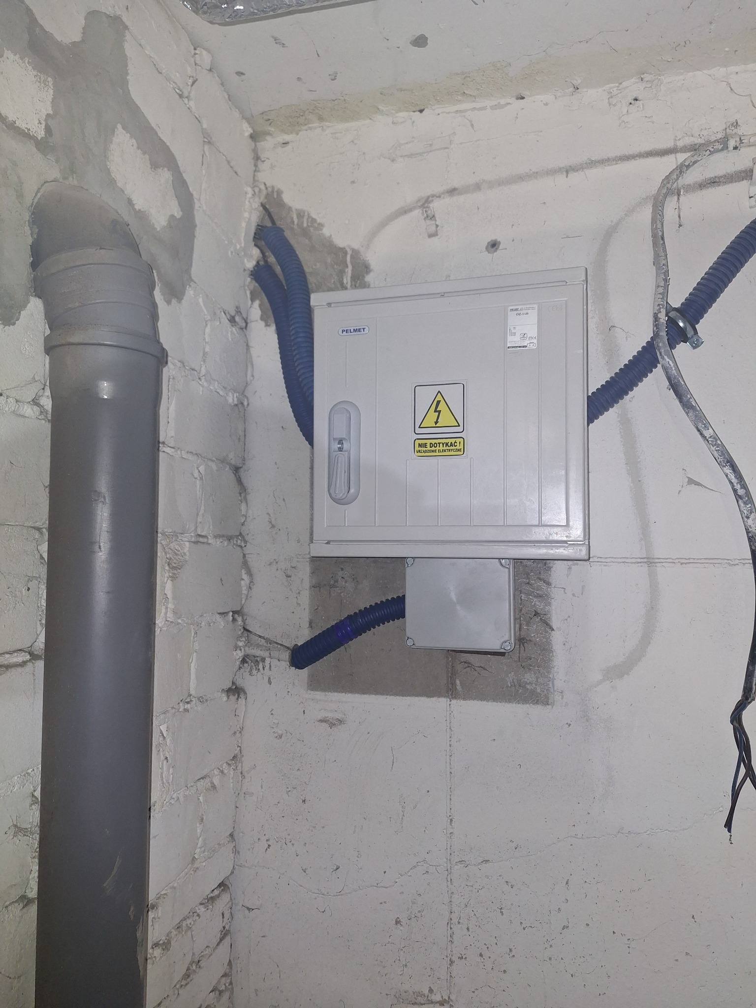 Szara skrzynka elektryczna PELMET z żółtym znakiem ostrzegawczym na ścianie z instalacją elektryczną w nieotynkowanym pomieszczeniu.