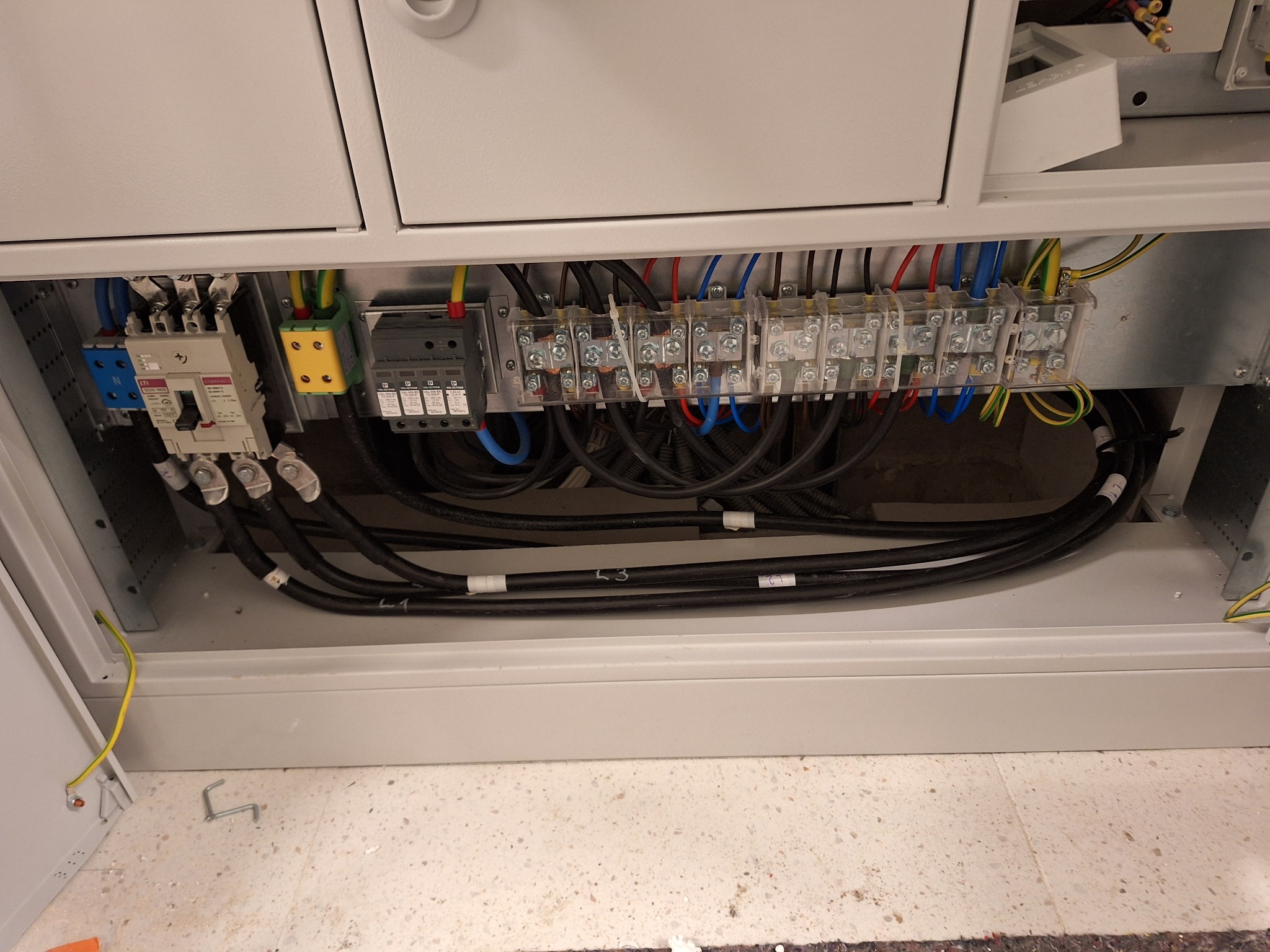 Wnętrze szafy elektrycznej z bezpiecznikami, przewodami oznaczonymi etykietami, listwami zaciskowymi i elementami instalacji, widoczne połączenia i kable.