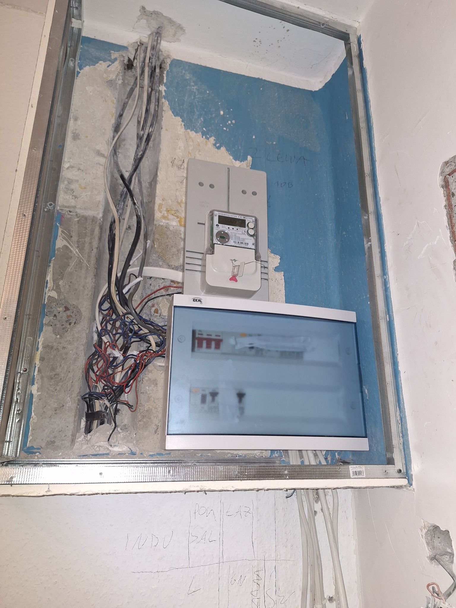 Wnęka z zamontowaną skrzynką elektryczną, licznikiem i przewodami. Widoczne kable w ścianie i metalowa rama. Ściana w surowym stanie, z widocznymi śladami po kuciu.