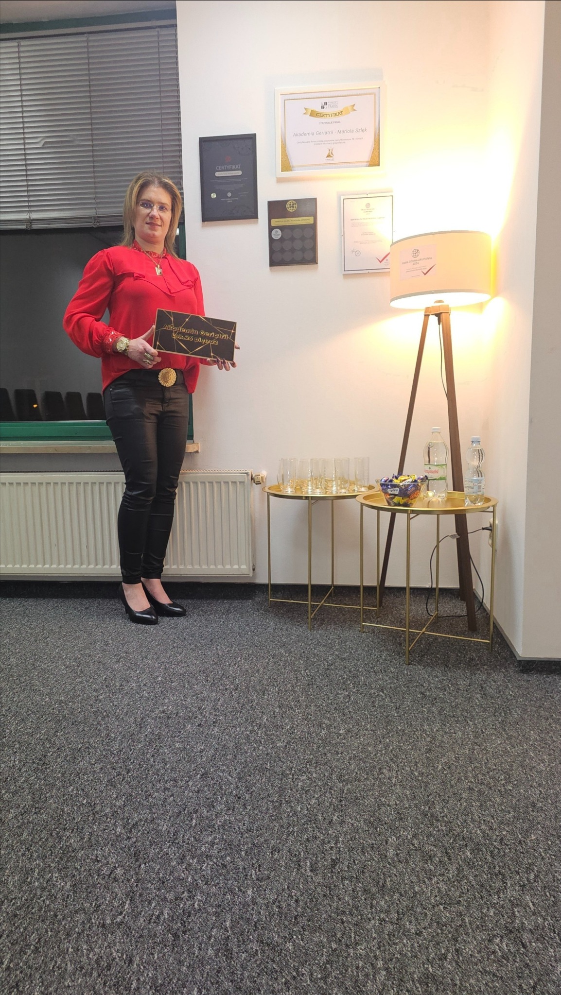 Kobieta w czerwonej bluzce i czarnych spodniach trzyma tabliczkę z napisem 'Akademia Geriatrii Lekarz Plus'. Na ścianie wiszą certyfikaty, a obok stoją stoliki z kieliszkami i wodą.