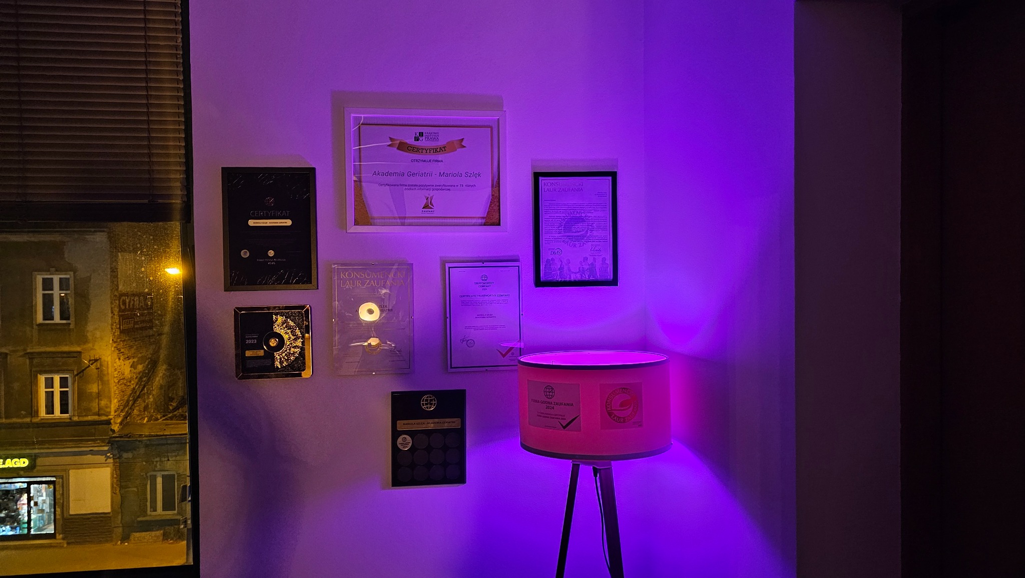 Ściana oświetlona fioletowym światłem, na której wiszą certyfikaty i nagrody, widoczne okno z widokiem na budynek.