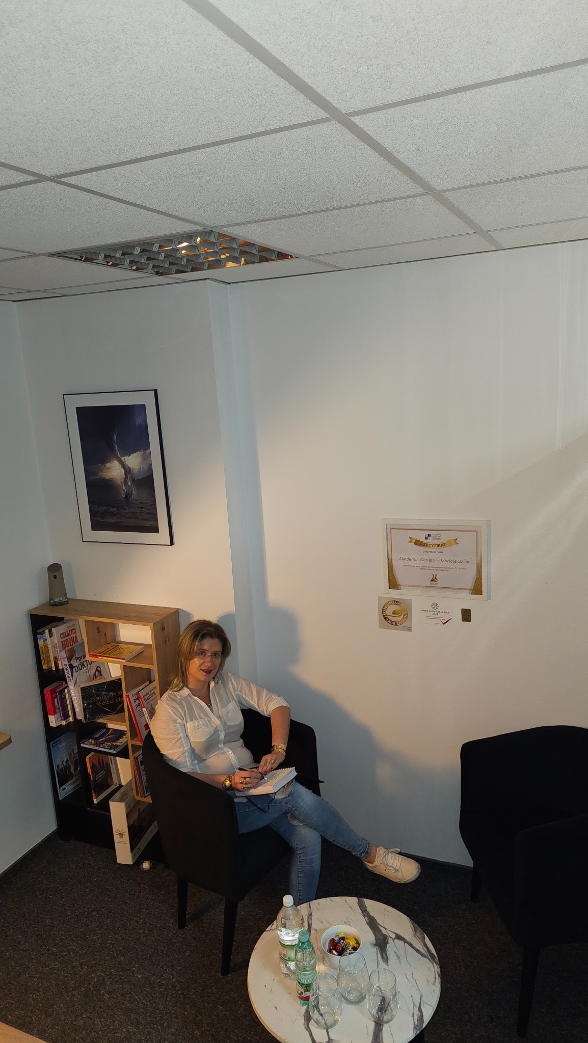 Kobieta w białej koszuli siedzi w czarnym fotelu, pisząc w notatniku. Na ścianie wisi certyfikat Akademii Geriatrii Mariola Szłek. Na stoliku woda i cukierki.