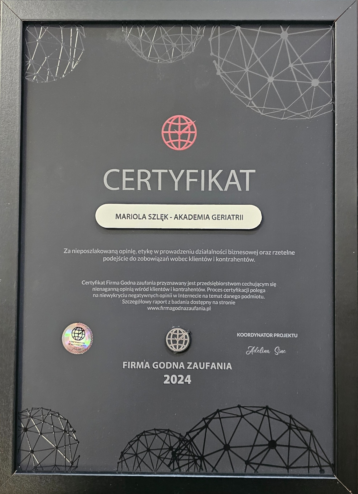 Oprawiony certyfikat 'Firma Godna Zaufania 2024' dla Marioli Szlęk - Akademia Geriatrii, przyznany za nieposzlakowaną opinię i etykę w biznesie, z hologramem potwierdzającym autentyczność...
