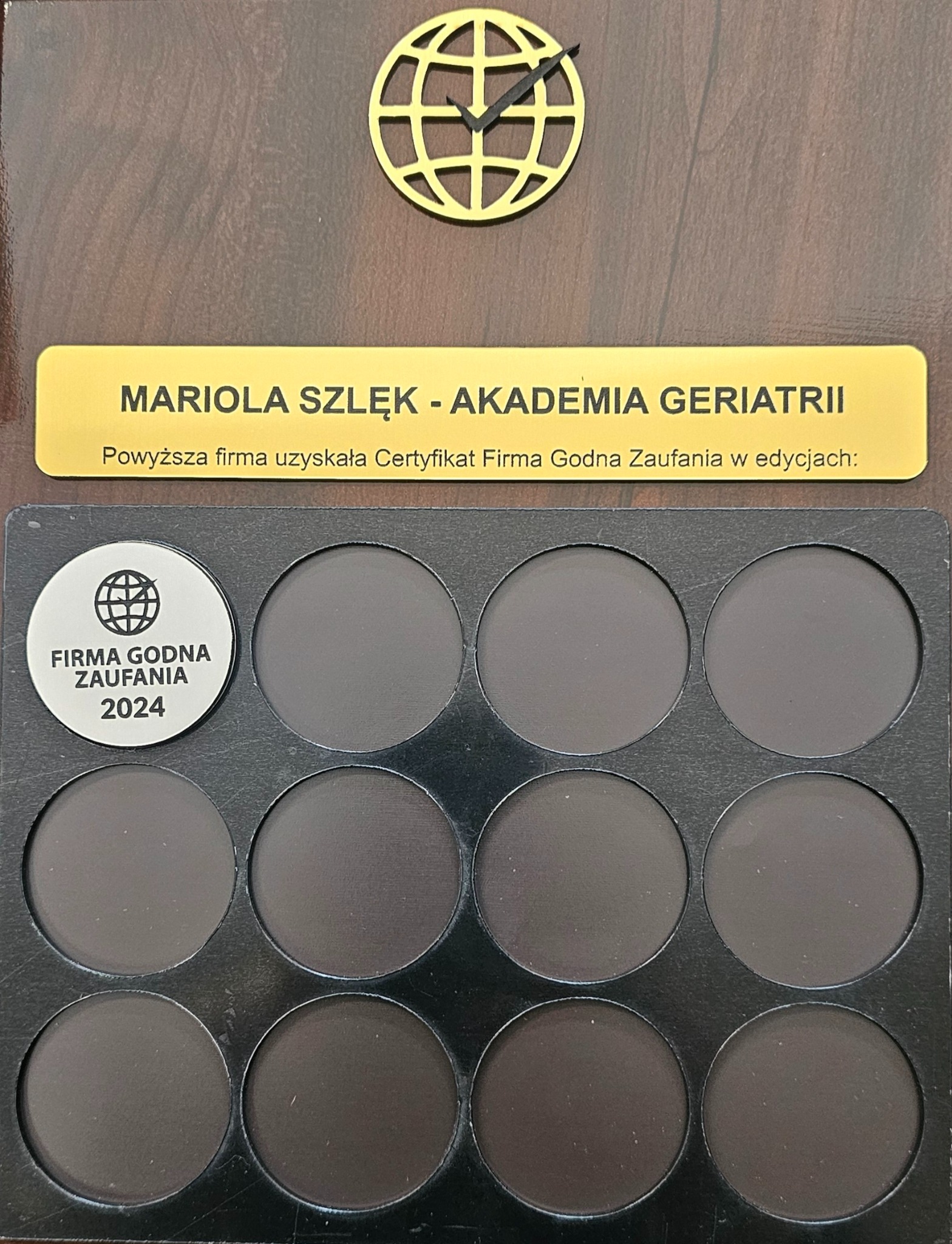 Tablica z certyfikatem 'Firma Godna Zaufania 2024' dla Marioli Szlęk - Akademia Geriatrii, prezentowana na drewnianym tle z logo firmy w formie globu z zaznaczoną godziną.