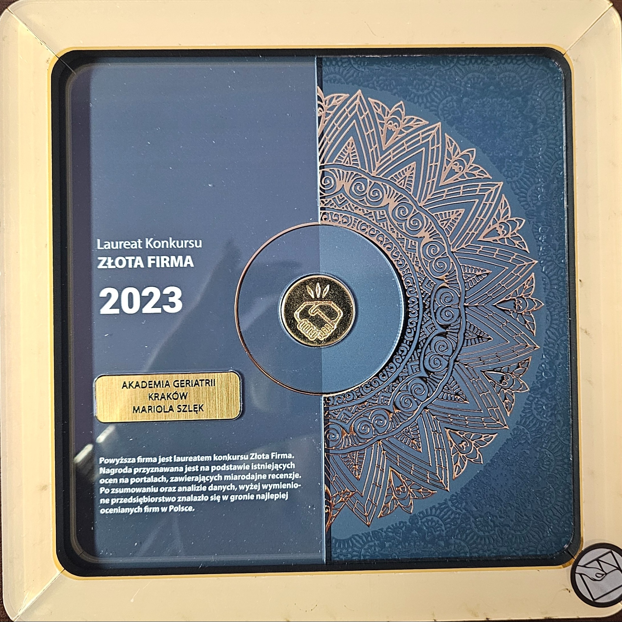 Nagroda Złota Firma 2023 dla Akademii Geriatrii Kraków Mariola Szlęk, prezentowana w ramce na tle dekoracyjnym z motywem mandali i złotą monetą z symbolem uścisku dłoni.