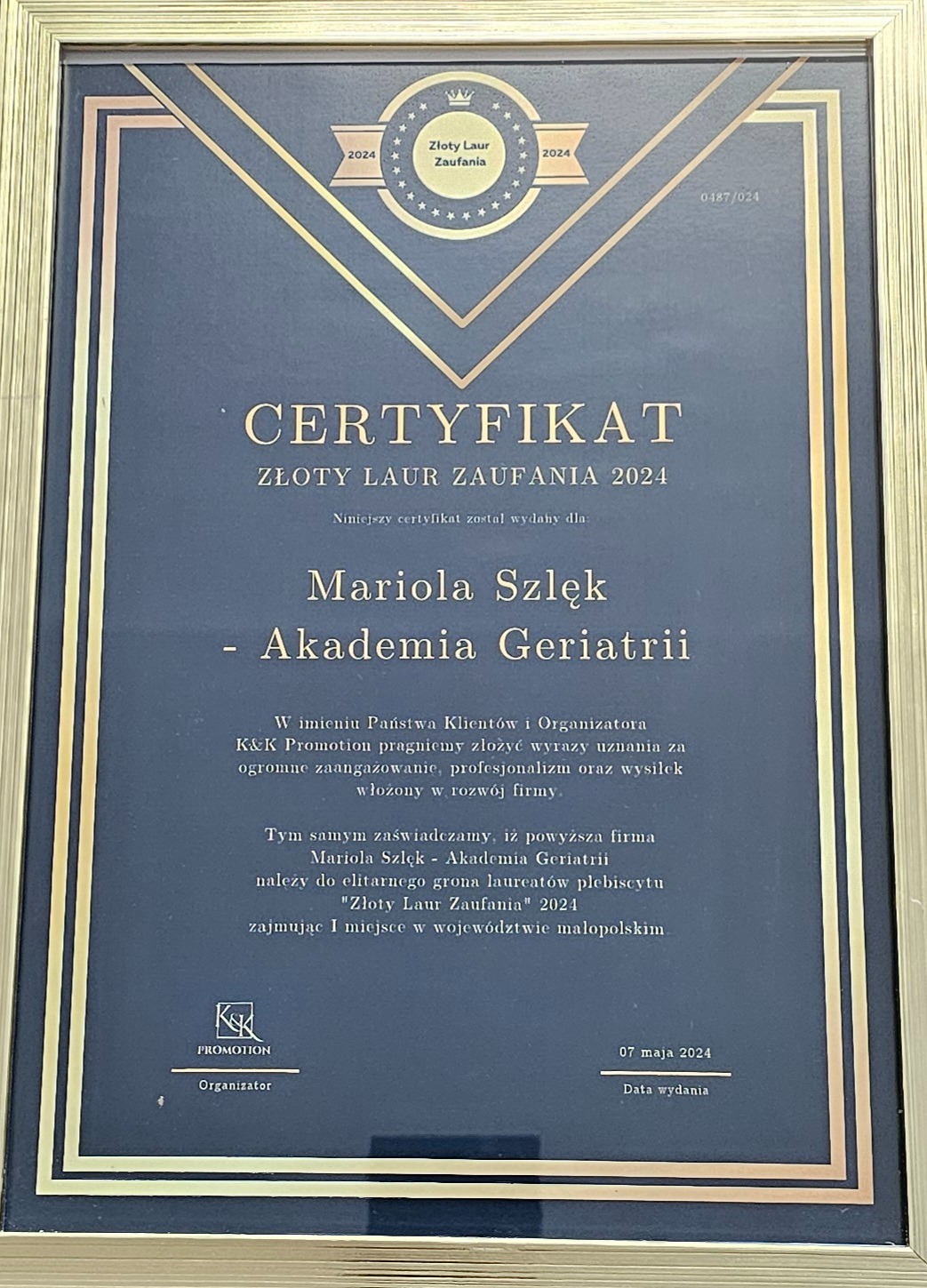 Certyfikat 'Złoty Laur Zaufania 2024' dla Marioli Szlęk - Akademia Geriatrii z Krakowa, oprawiony w złotą ramę, potwierdzający wysokie zaangażowanie i profesjonalizm firmy.
