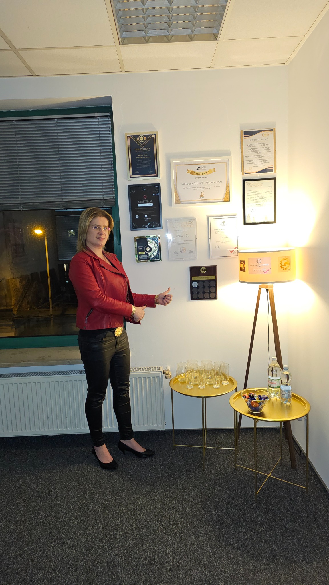 Kobieta w czerwonej kurtce i czarnych spodniach pozuje na tle ściany z certyfikatami i listami gratulacyjnymi, obok stolików z szklankami i butelkami wody.