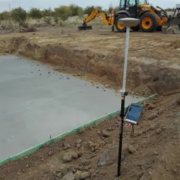 Wykonana płyta fundamentowa z użyciem niwelatora GPS na tyczce, w tle koparka JCB. Widoczne szalunki i wykop. Prace ziemne i geodezyjne na budowie.
