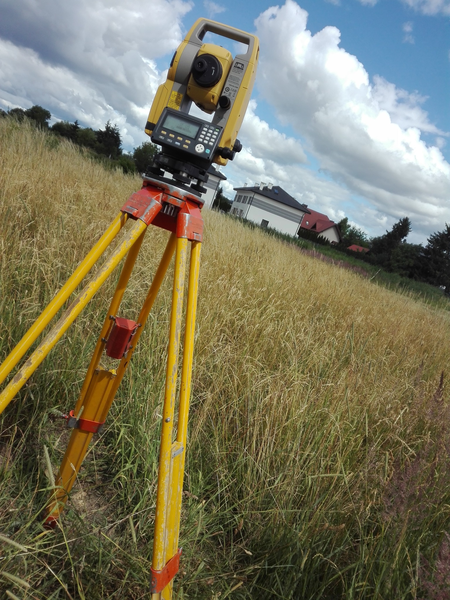 Żółty teodolit ustawiony na trójnogu na polu z wysoką trawą, w tle budynki mieszkalne i pochmurne niebo.