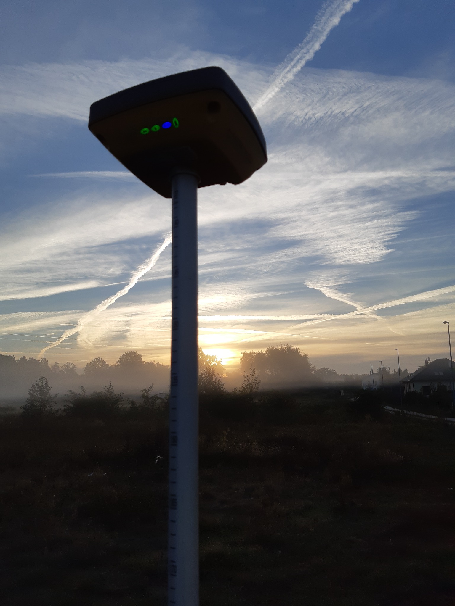 Profesjonalny odbiornik GNSS na tyczce geodezyjnej, w tle mglista łąka o świcie i niebo z smugami kondensacyjnymi.