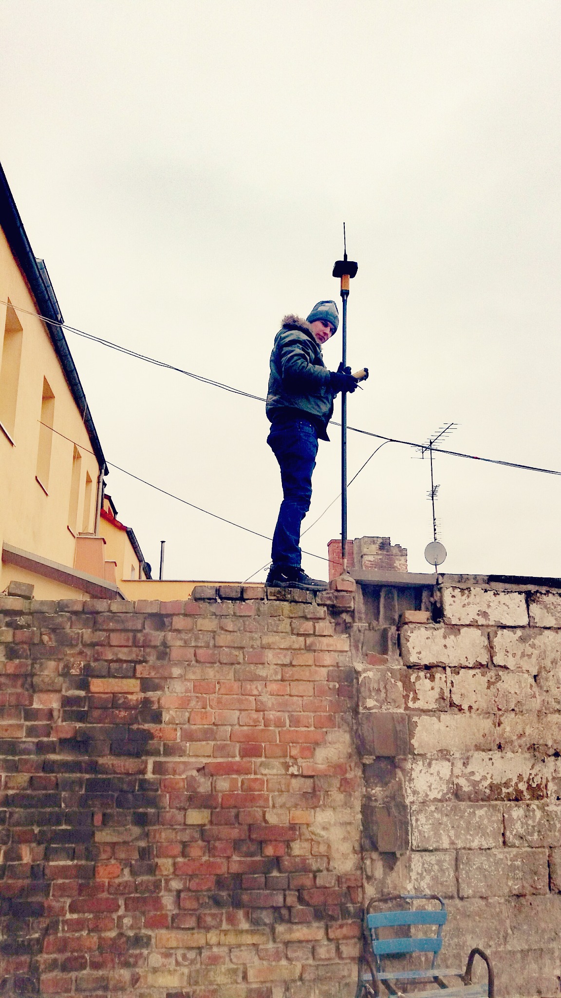Osoba w kurtce i czapce stoi na murze z cegły, trzymając tyczkę geodezyjną z anteną, budynek w tle.