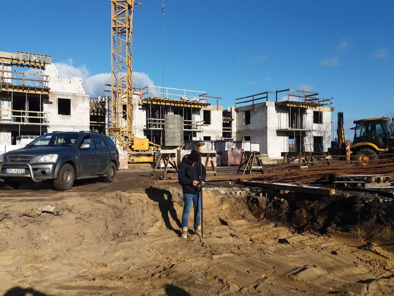 Pracownik w czapce dokonuje pomiarów geodezyjnych na placu budowy z użyciem niwelatora. W tle budynki w stanie surowym, dźwig i samochód.