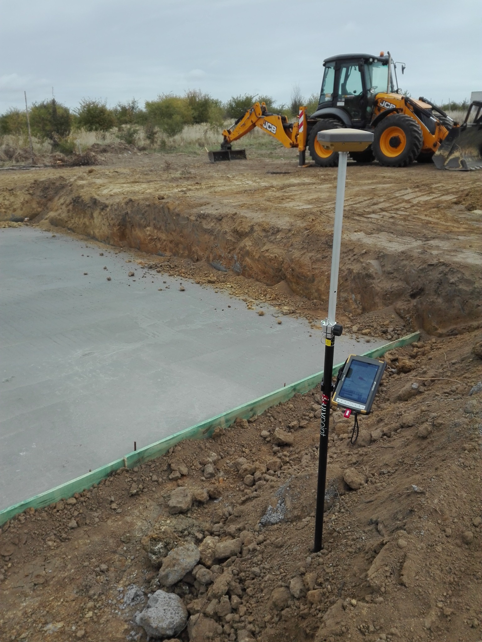 Wykonana płyta fundamentowa z użyciem niwelatora GPS na tyczce, w tle koparka JCB. Widoczne szalunki i wykop. Prace ziemne i geodezyjne na budowie.