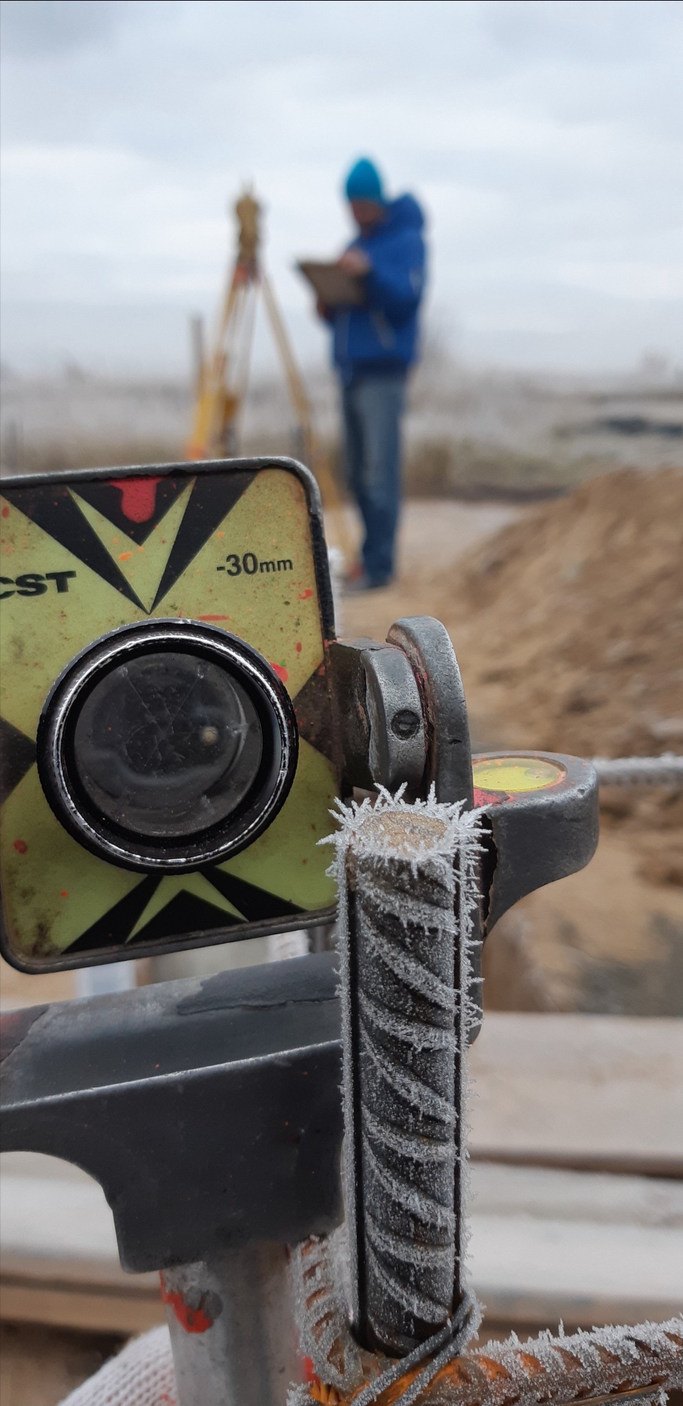 Przymrożony reflektor geodezyjny CST na tle pracownika z niwelatorem. Detal pokryty szronem, w tle ziemia i pochmurne niebo.