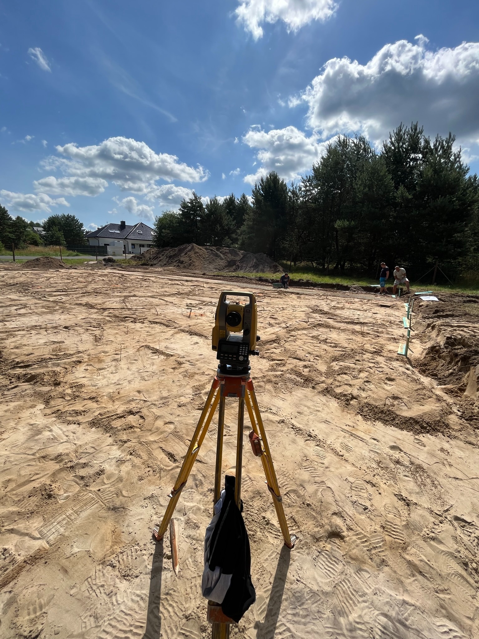 Teren budowy z niwelatorem na statywie w centrum kadru. W tle widoczny dom, drzewa i pracownicy. Błękitne niebo z chmurami. Ślady opon i butów na piasku.
