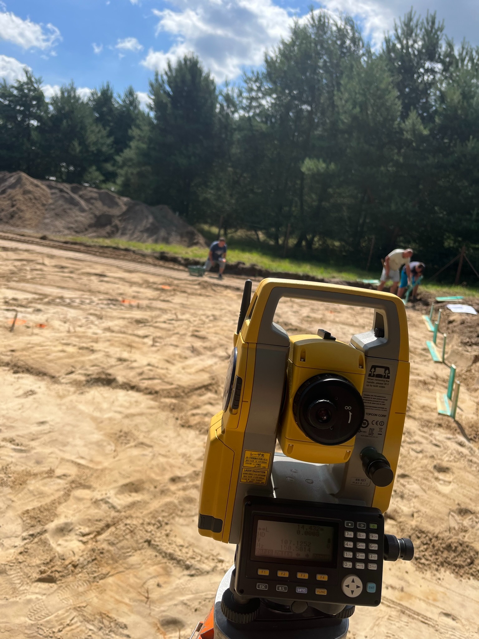 Żółty teodolit na placu budowy z ekipą w tle, pomiary geodezyjne w toku. Widok na teren z drzewami i błękitnym niebem.