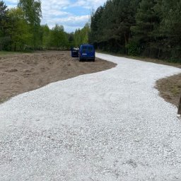 RDT Bud - Wysypana, biała, kamienista droga dojazdowa na działce z otwartymi drzwiami niebieskiego vana w tle i drewnianym płotem po prawej stronie.