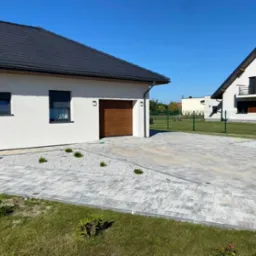 Nowoczesny podjazd z szarej kostki brukowej ułożonej we wzór, z białym żwirem i niskimi roślinami ozdobnymi, na tle białego domu z brązową bramą garażową i zadbanego trawnika.