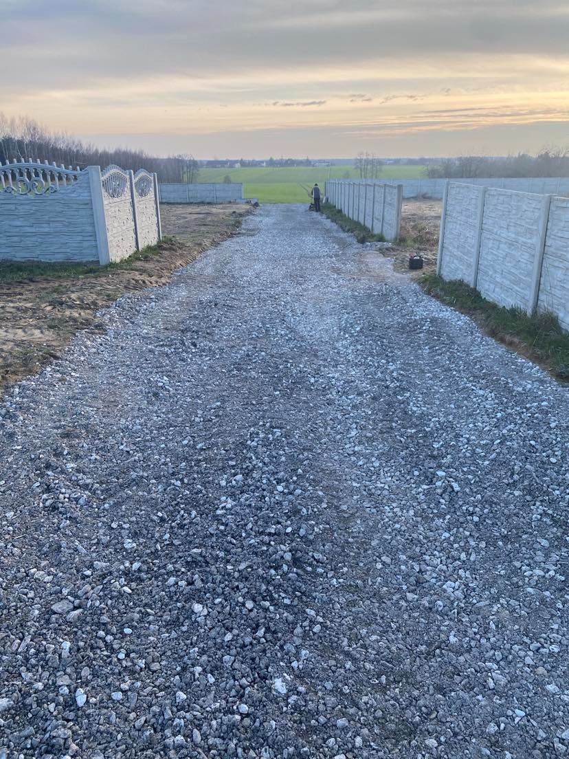 Długa, wysypana szutrem droga wzdłuż betonowego ogrodzenia panelowego, widok na oddalony krajobraz z polami i lasem, osoba z maszyną w tle.