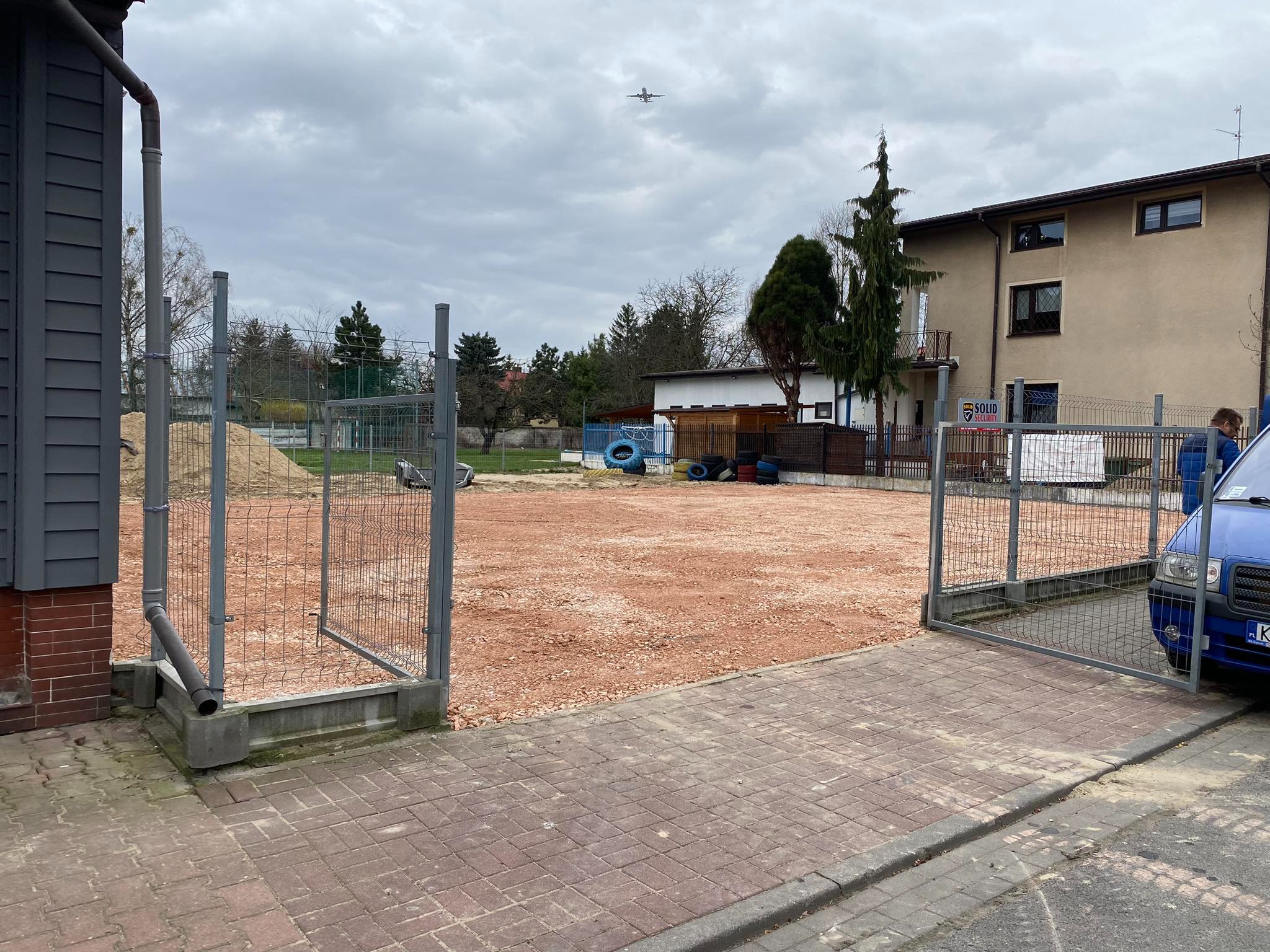 Teren przygotowany pod budowę z widoczną czerwoną podsypką, otoczony metalową siatką i budynkami, z samolotem na pochmurnym niebie