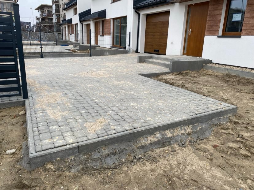 Nowo ułożona, szara kostka brukowa przed szeregiem domów jednorodzinnych w trakcie wykończenia, widoczny piasek na powierzchni i niezabezpieczone krawędzie.