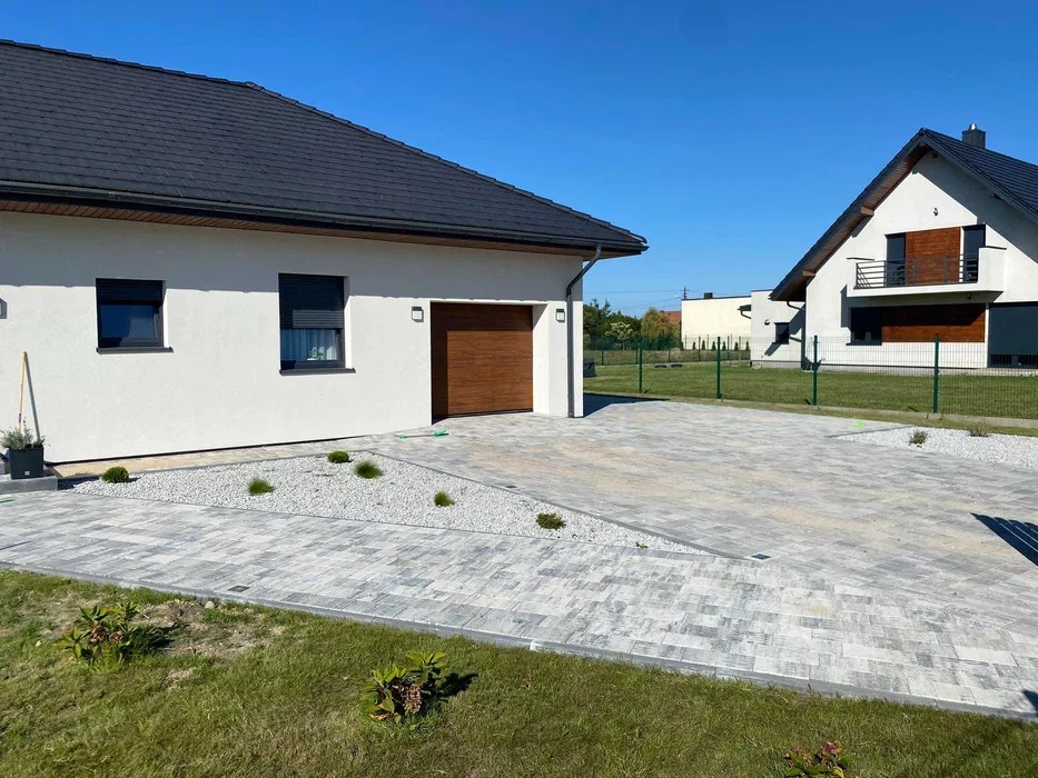 Nowoczesny podjazd z szarej kostki brukowej ułożonej we wzór, z białym żwirem i niskimi roślinami ozdobnymi, na tle białego domu z brązową bramą garażową i zadbanego trawnika.