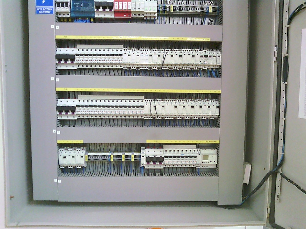Otwarta szafa sterownicza z widocznymi bezpiecznikami, stycznikami i okablowaniem, widoczny napis 'Wyłącznik Główny'.