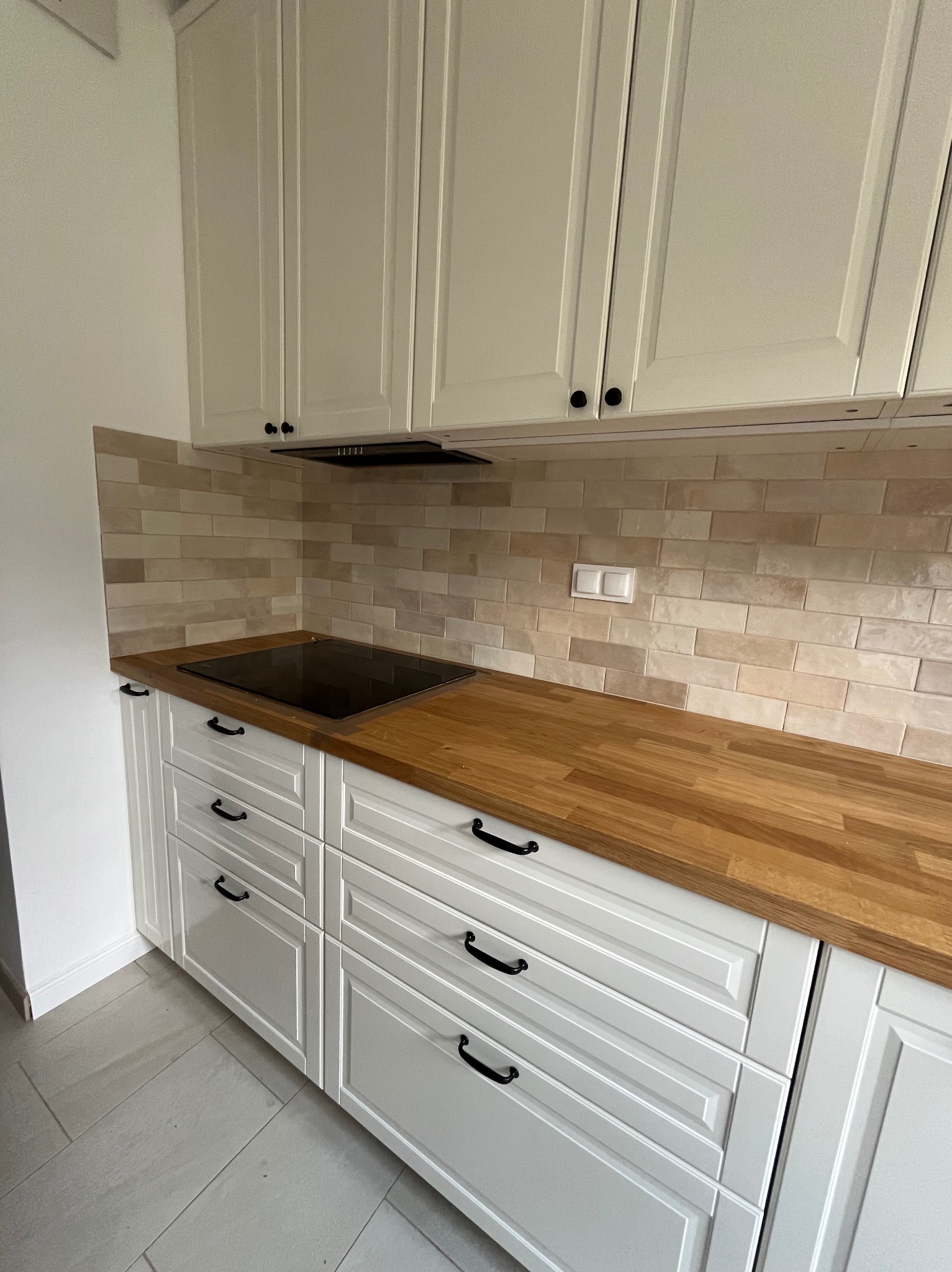 Jasna kuchnia z białymi szafkami z frezowanymi frontami, drewnianym blatem i beżowymi płytkami na ścianie nad blatem.