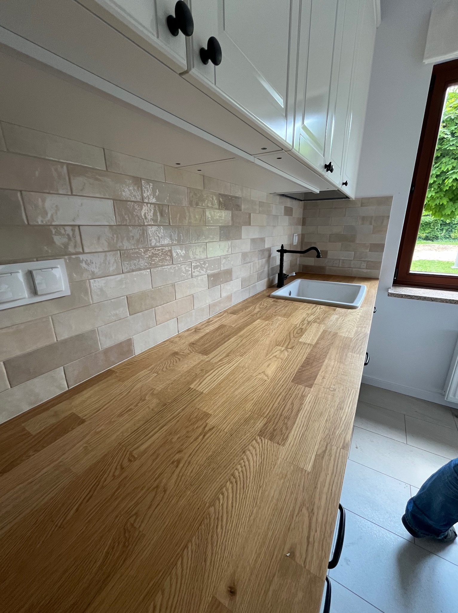 Jasna kuchnia z drewnianym blatem, beżowymi płytkami imitującymi cegłę i białymi szafkami wiszącymi z czarnymi uchwytami, widok na okno z zielenią na zewnątrz.