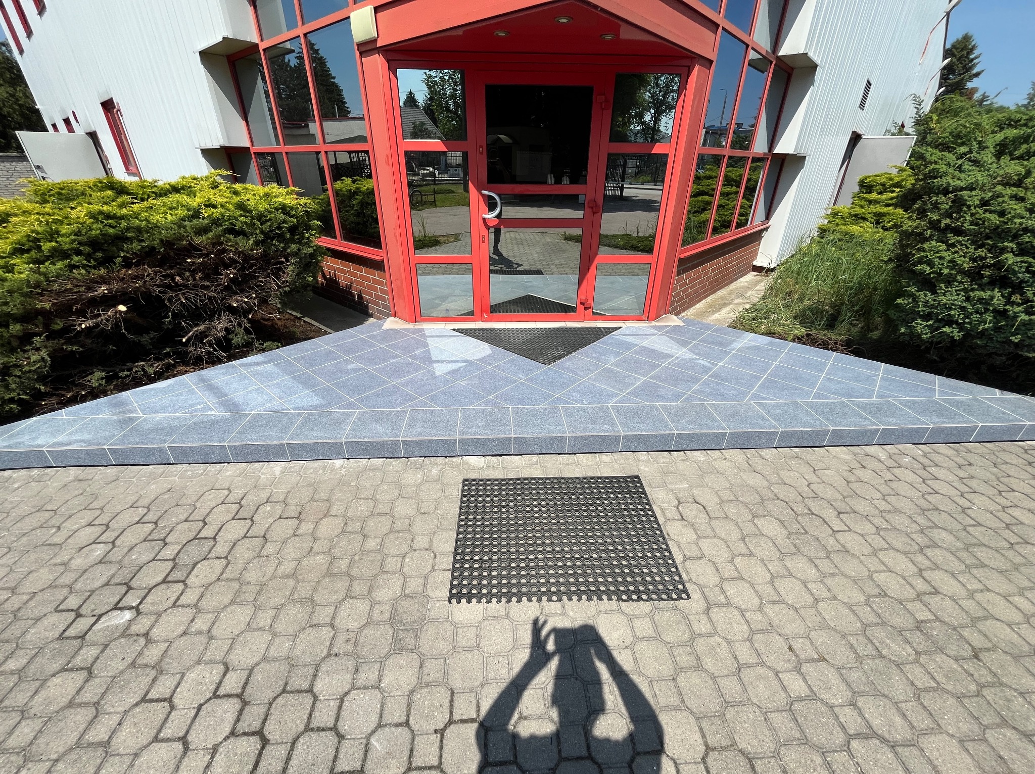 Wejście do budynku z czerwonymi ramami okiennymi i drzwiami, wyłożone niebiesko-szarą płytką ceramiczną, z widoczną wycieraczką i cieniem osoby robiącej zdjęcie.