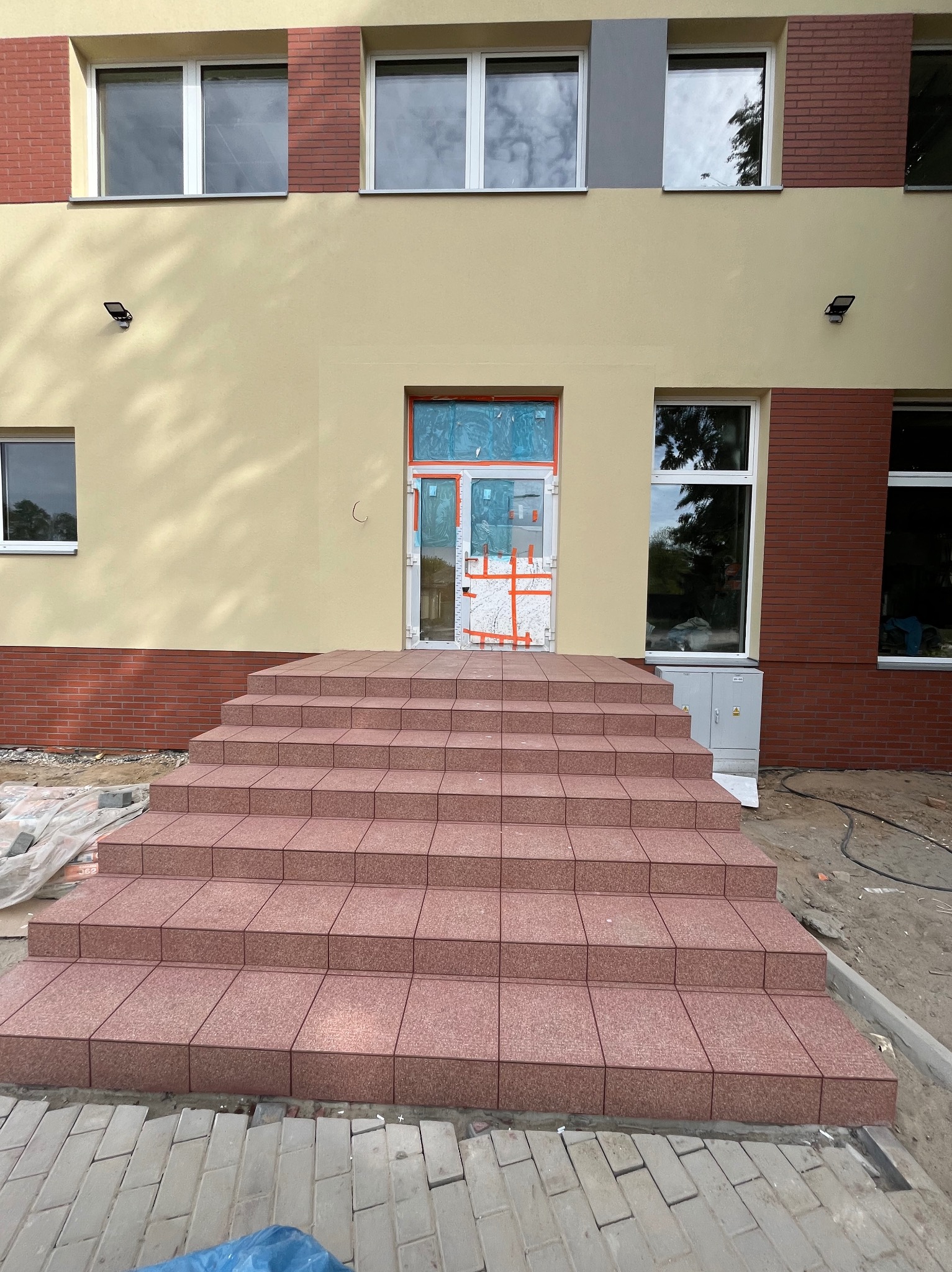 Nowe schody wejściowe wyłożone czerwoną terakotą, prowadzące do budynku z beżową elewacją i ceglanymi elementami, drzwi wejściowe zabezpieczone folią ochronną.