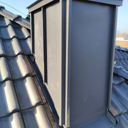 Dek-Id - Szary, metalowy komin wentylacyjny na dachu pokrytym ciemną dachówką ceramiczną, widoczne obróbki blacharskie u podstawy komina, błękitne niebo w tle.