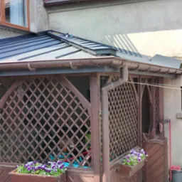 Nowy, blaszany dach altany z brązowymi rynnami, kratą i skrzynkami z bratkami, na tle starego domu z eternitem.