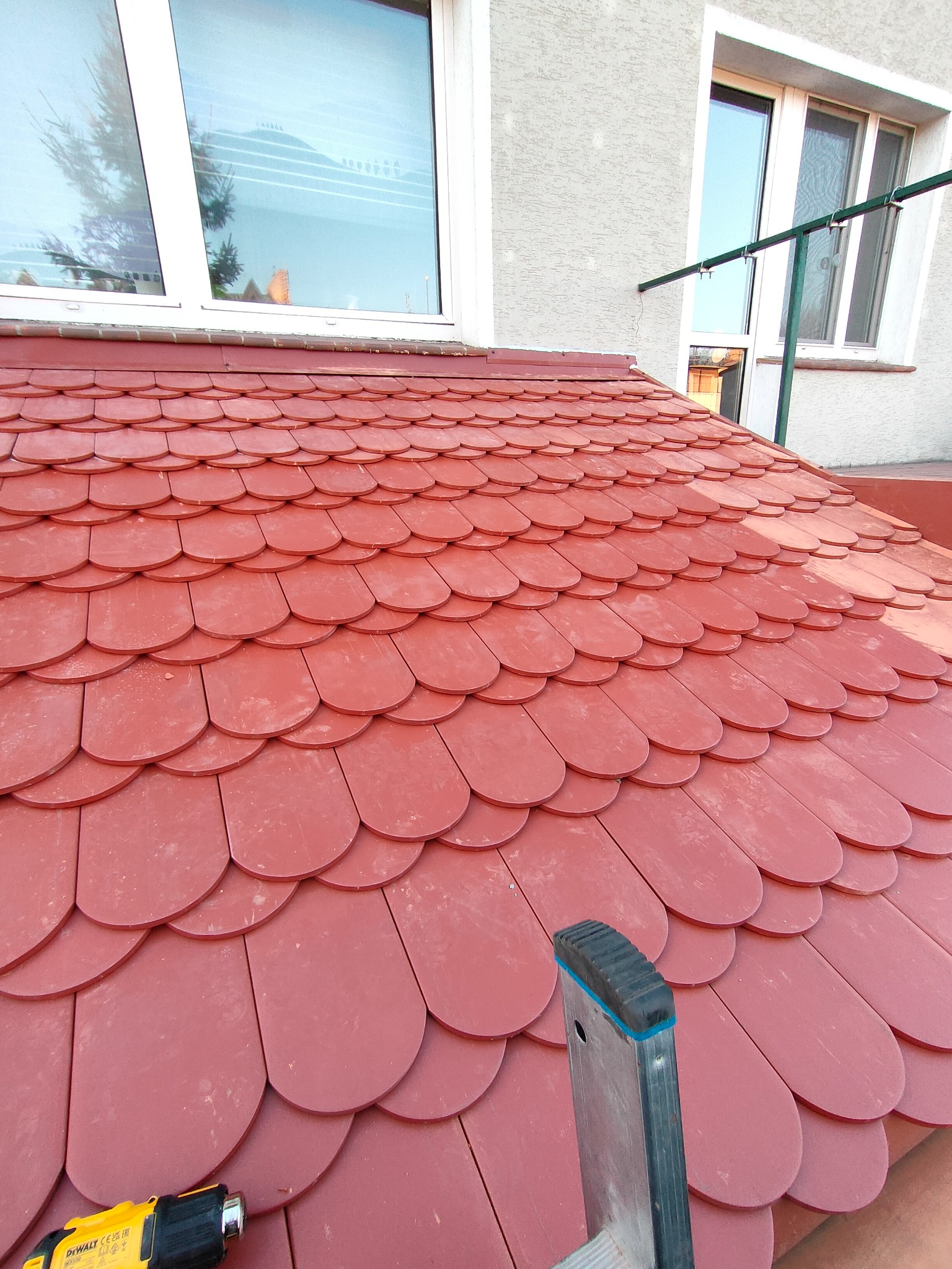 Czerwony dach pokryty dachówką bitumiczną w kształcie bobrówki, widoczne okna na poddaszu i fragment drabiny oraz opalarka dekarska