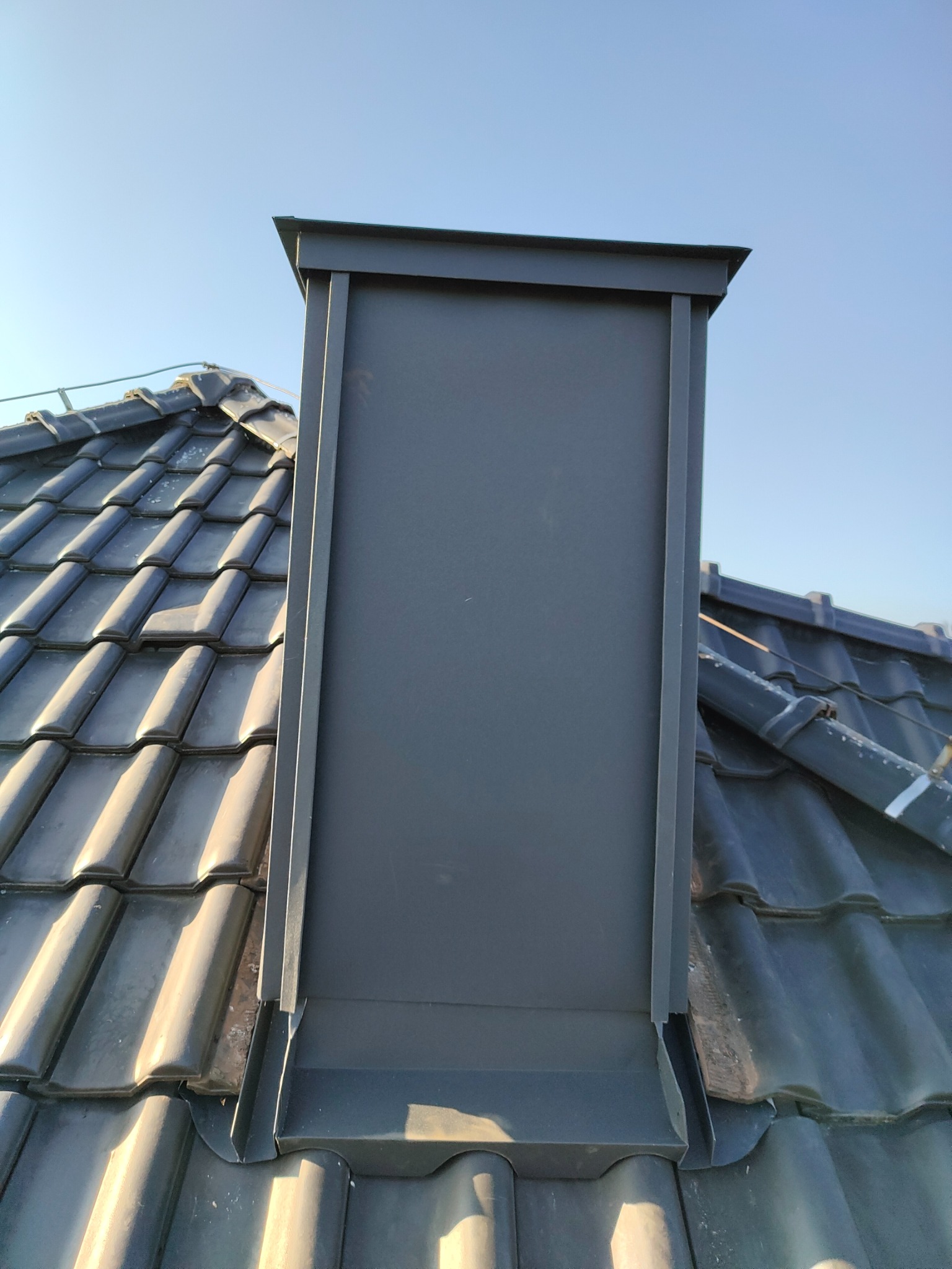Szary komin wentylacyjny z blachy na dachu pokrytym ciemną dachówką, widok z dołu na tle błękitnego nieba.