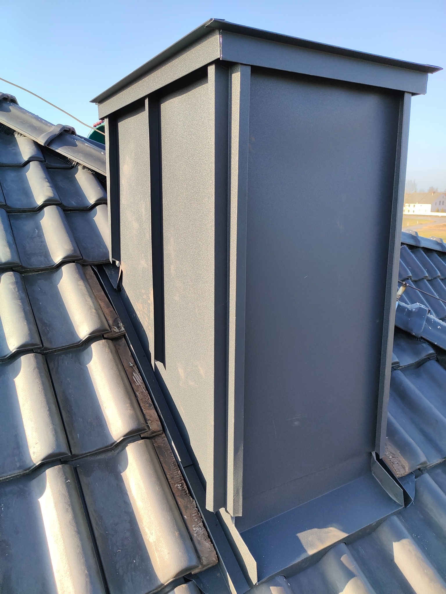 Szary, metalowy komin wentylacyjny na dachu pokrytym ciemną dachówką ceramiczną, widoczne obróbki blacharskie u podstawy komina, błękitne niebo w tle.