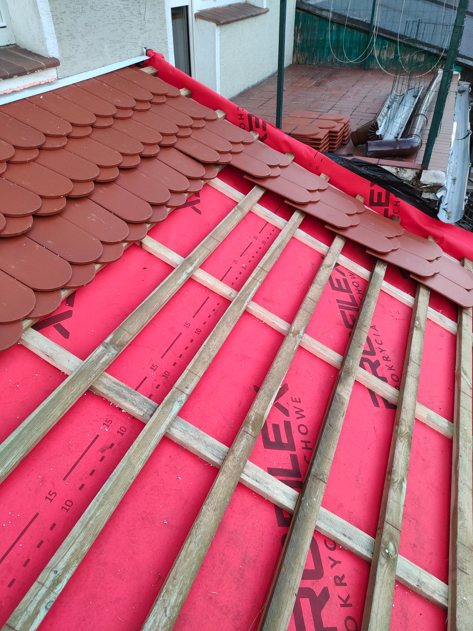 Montaż brązowej dachówki karpiówki na łatach drewnianych pokrytych czerwoną membraną dachową, widoczne elementy więźby dachowej i fragment rynny.