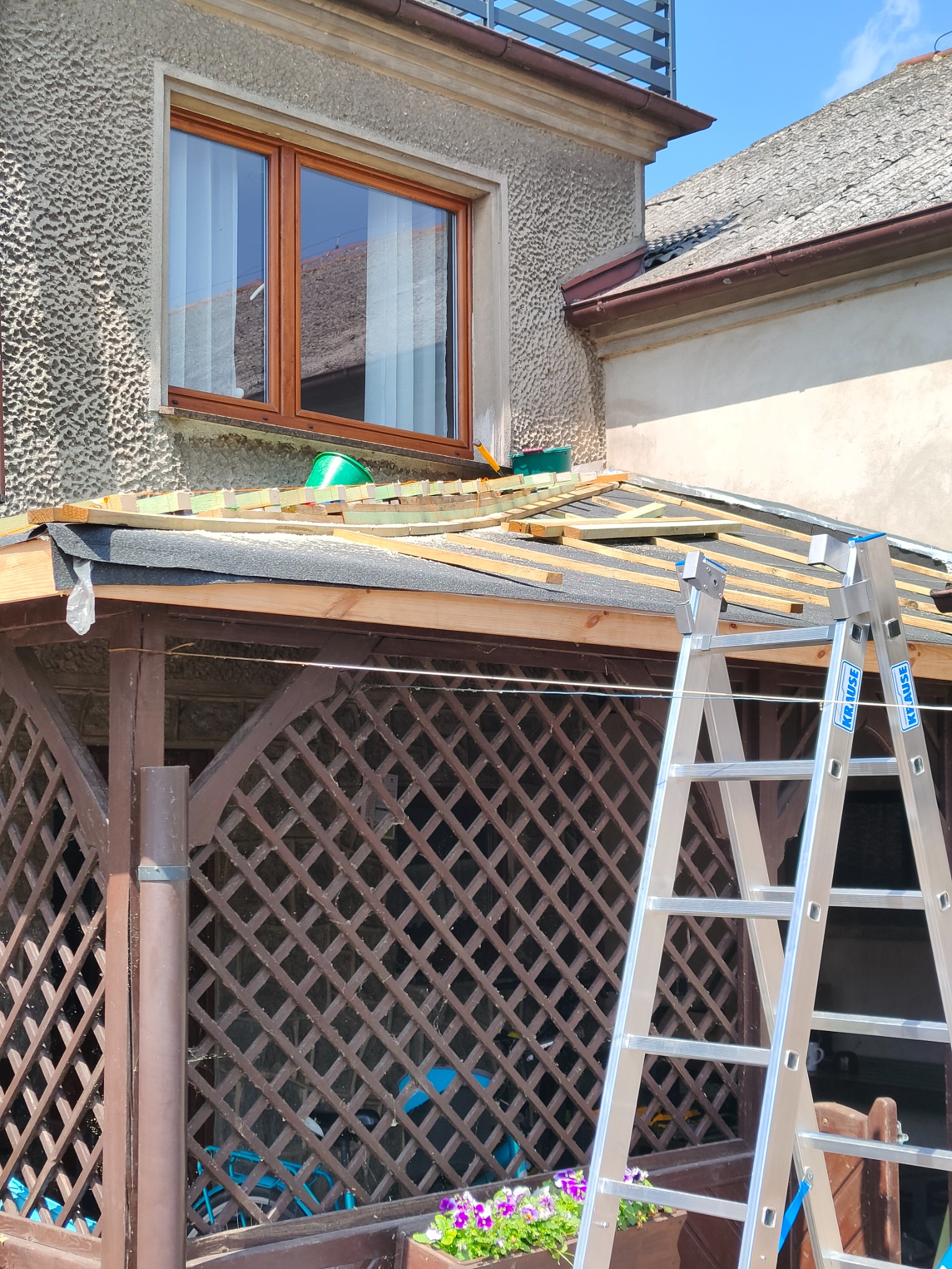 Konstrukcja dachu w trakcie budowy z widocznymi łatami, membraną i drabiną opartą o drewnianą altanę.