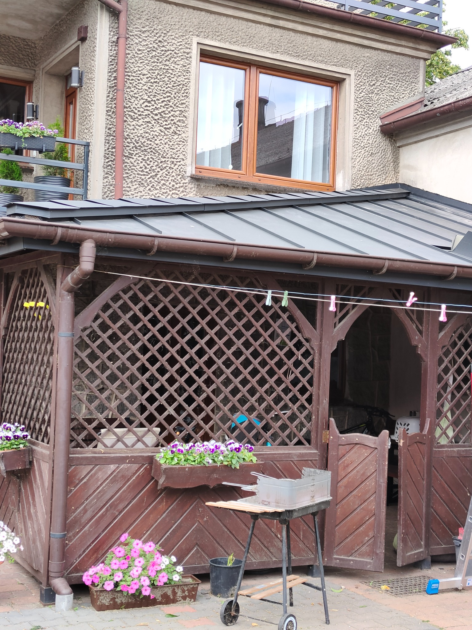 Zadaszona altana z drewnianą kratą, metalowym dachem i rynnami w kolorze brązowym, przed nią grill na kółkach i kwiaty w donicach, w tle budynek mieszkalny z oknami.