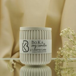 Weronika Jędrzejak - Biała, prążkowana świeca sojowa z etykietą 'BO.KA soy candle Livorno Lady' na lustrzanym podłożu, ozdobiona gałązką gipsówki, na tle drapowanej beżowej tkaniny.