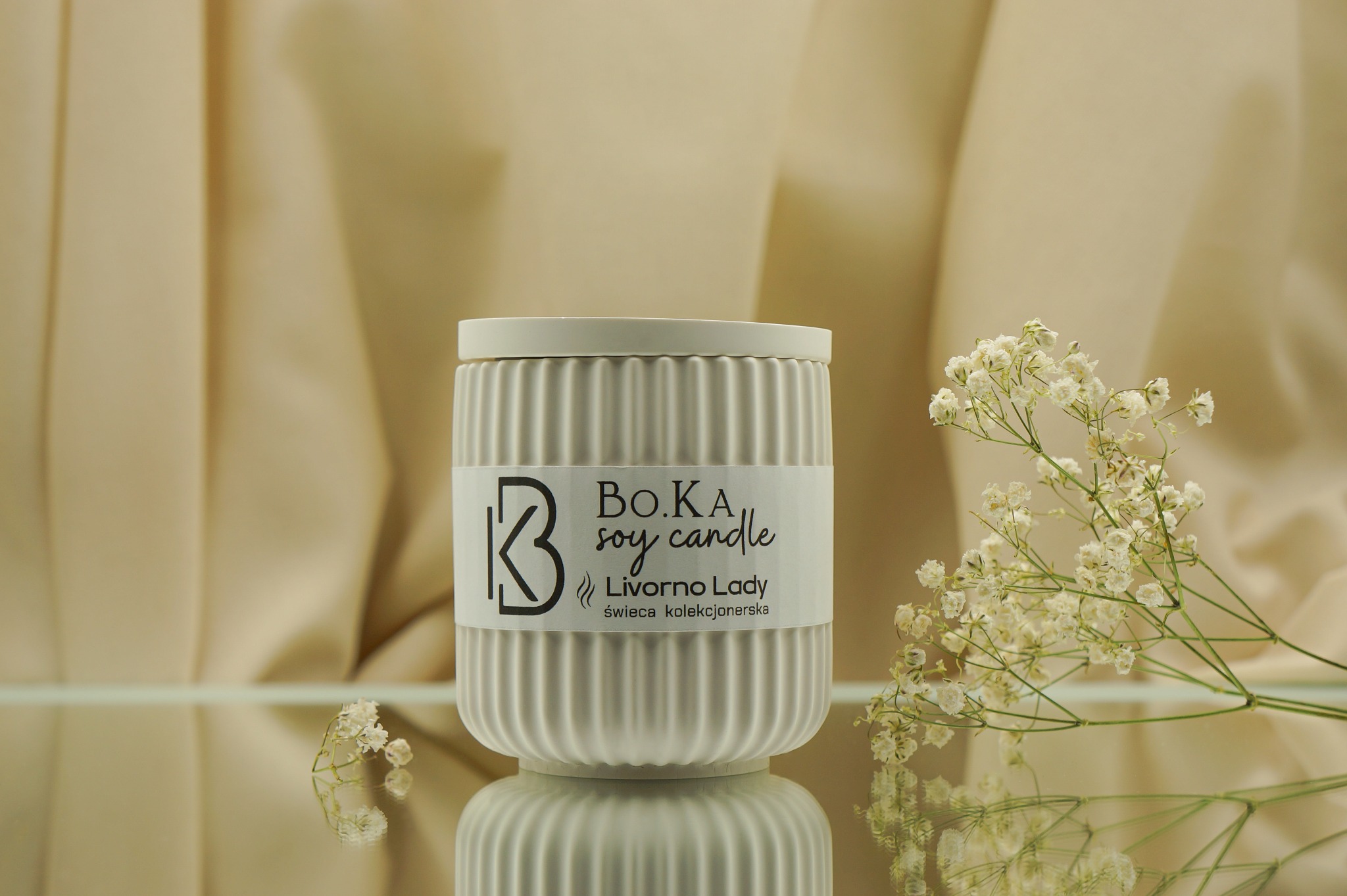 Biała, prążkowana świeca sojowa z etykietą 'BO.KA soy candle Livorno Lady' na lustrzanym podłożu, ozdobiona gałązką gipsówki, na tle drapowanej beżowej tkaniny.