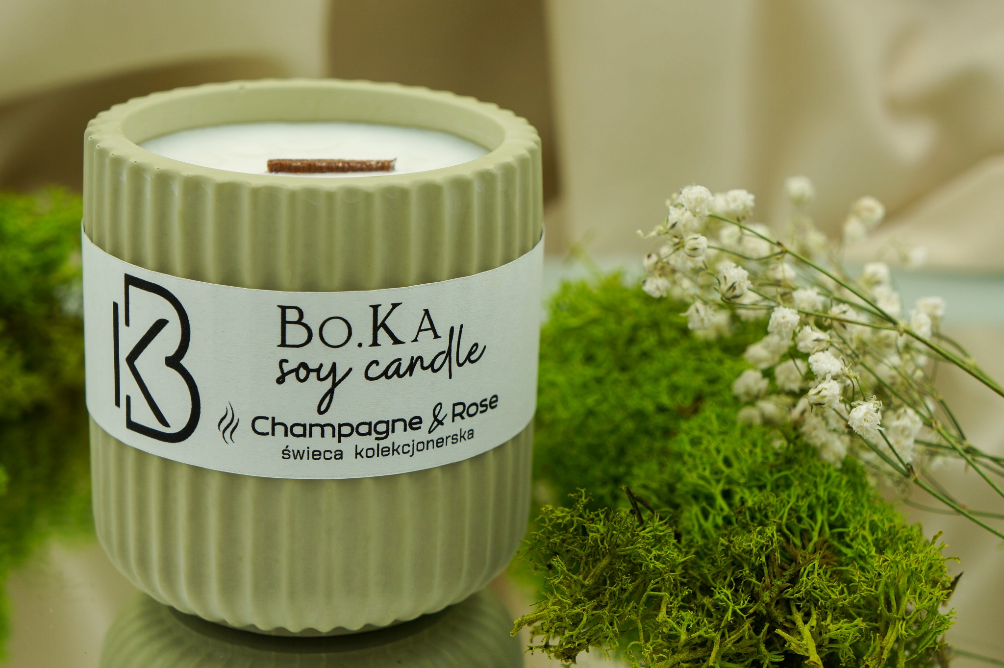Zdjęcie sojowej świecy kolekcjonerskiej 'Champagne & Rose' w minimalistycznym, żłobionym, oliwkowym pojemniku, z drewnianym knotem, na lustrzanej powierzchni, udekorowanej mchem i drobnymi białymi...