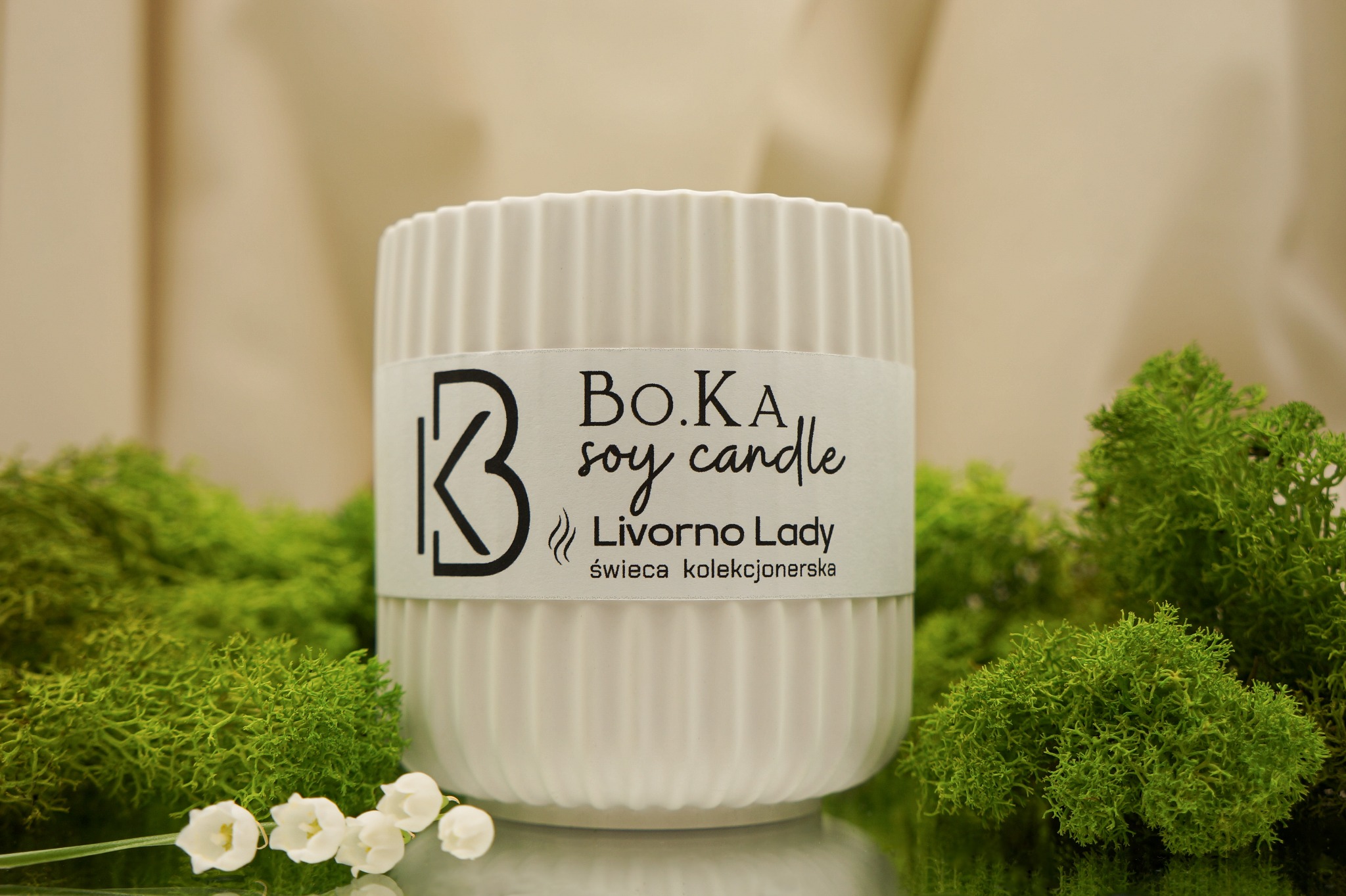 Biała, żłobkowana świeca sojowa B.O.K.A Livorno Lady, otoczona zielonym mchem i konwaliami na błyszczącym podłożu.