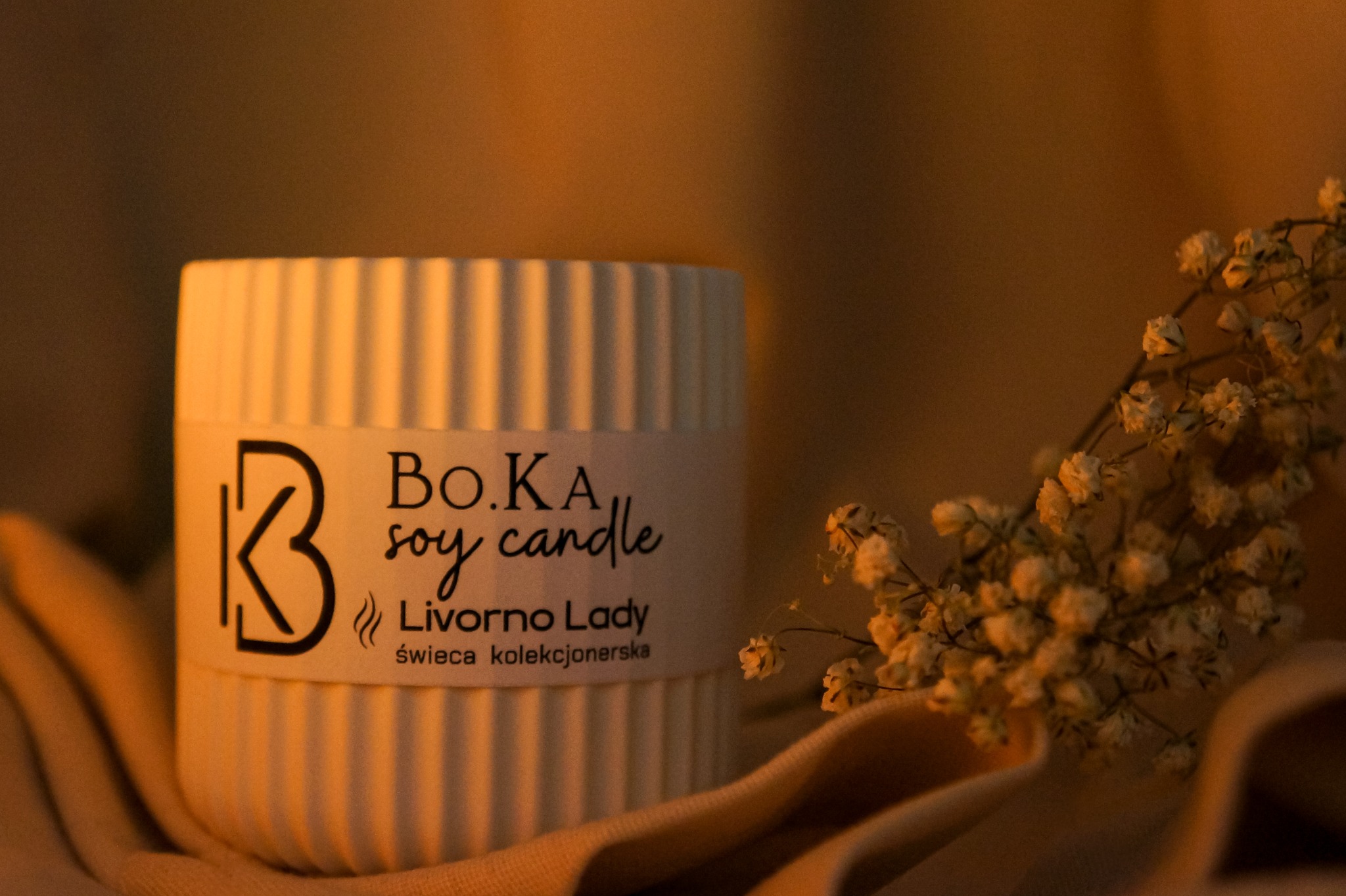 Beżowa świeca sojowa Bo.KA Livorno Lady z minimalistycznym logo i napisem, otoczona delikatnymi, zasuszonymi kwiatami na miękkiej, drapowanej tkaninie, sfotografowana w ciepłym, nastrojowym świetle.