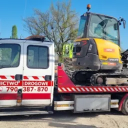 Laweta z żółto-szarą mini koparką Volvo EC35D na platformie, widoczny numer telefonu 790-387-870 i napis 24H Ratownictwo Drogowe na białej kabinie, w tle budynek z otwartą bramą.