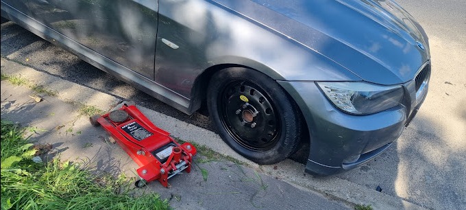 Szary samochód BMW z uszkodzonym przednim kołem, podnośnik hydrauliczny w kolorze czerwonym obok koła.