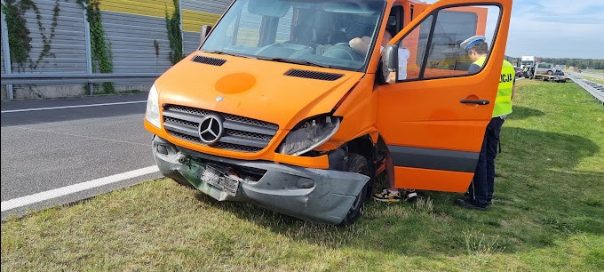 Pomarańczowy uszkodzony van Mercedes Sprinter na poboczu autostrady, z otwartymi drzwiami kierowcy, obok stoi policjant w żółtej kamizelce. Widoczne zniszczenia przodu pojazdu i zgnieciony zderzak.